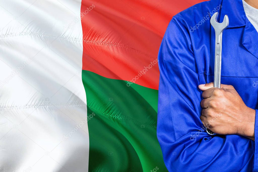 Malagasy Mechanic in blue uniform is holding wrench against waving Madagascar flag background. Crossed arms technician.