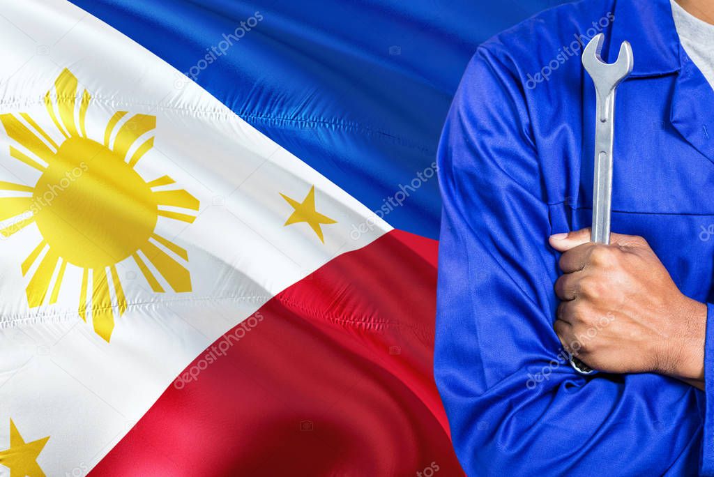 Filipino Mechanic in blue uniform is holding wrench against waving Philippines flag background. Crossed arms technician.
