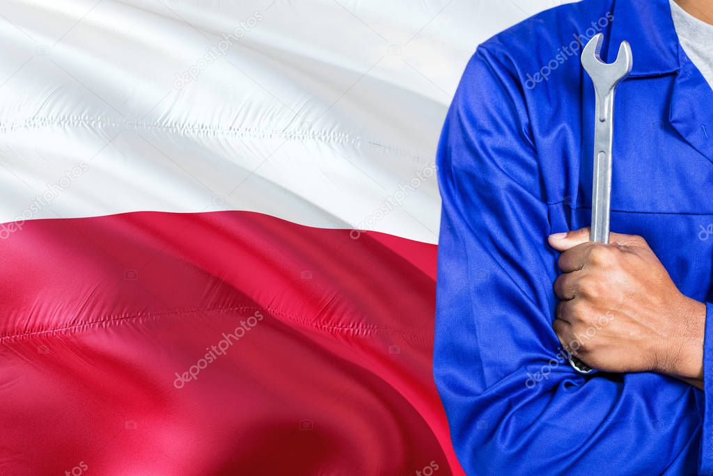 Polish Mechanic in blue uniform is holding wrench against waving Poland flag background. Crossed arms technician.