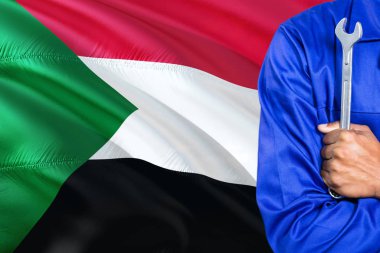 Mavi üniformalı sudan tamirci anahtarı sallayarak Sudan bayrağı arka planı tutuyor. Çapraz silah teknisyen.