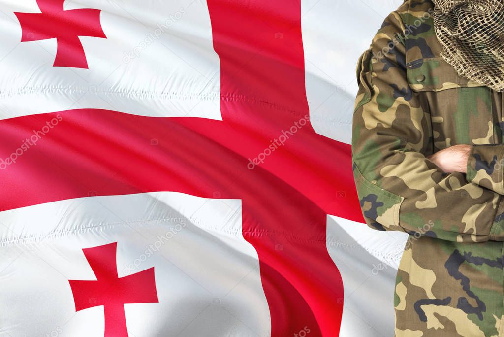 Crossed arms Georgian soldier with national waving flag on background - Georgia Military theme.