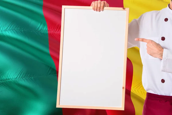 喀麦隆厨师拿着空白白板菜单在喀麦隆旗子背景 烹饪穿着统一的指向空间的文字 — 图库照片