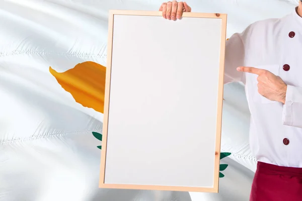 Cypriot Chef holding blank whiteboard menu on Cyprus flag background. Cook wearing uniform pointing space for text.