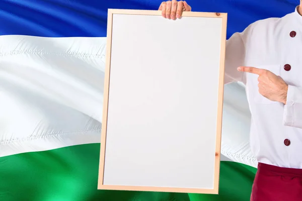 Chef holding blank whiteboard menu on Lesotho flag background. Cook wearing uniform pointing space for text.