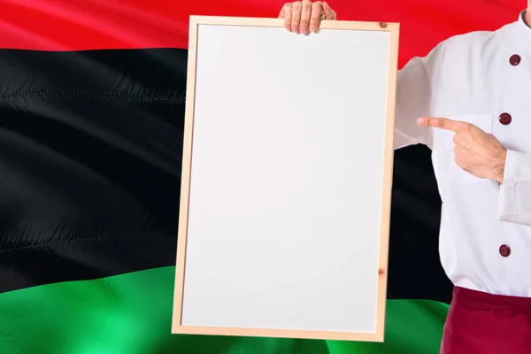 Libyan Chef holding blank whiteboard menu on Libya flag background. Cook wearing uniform pointing space for text.