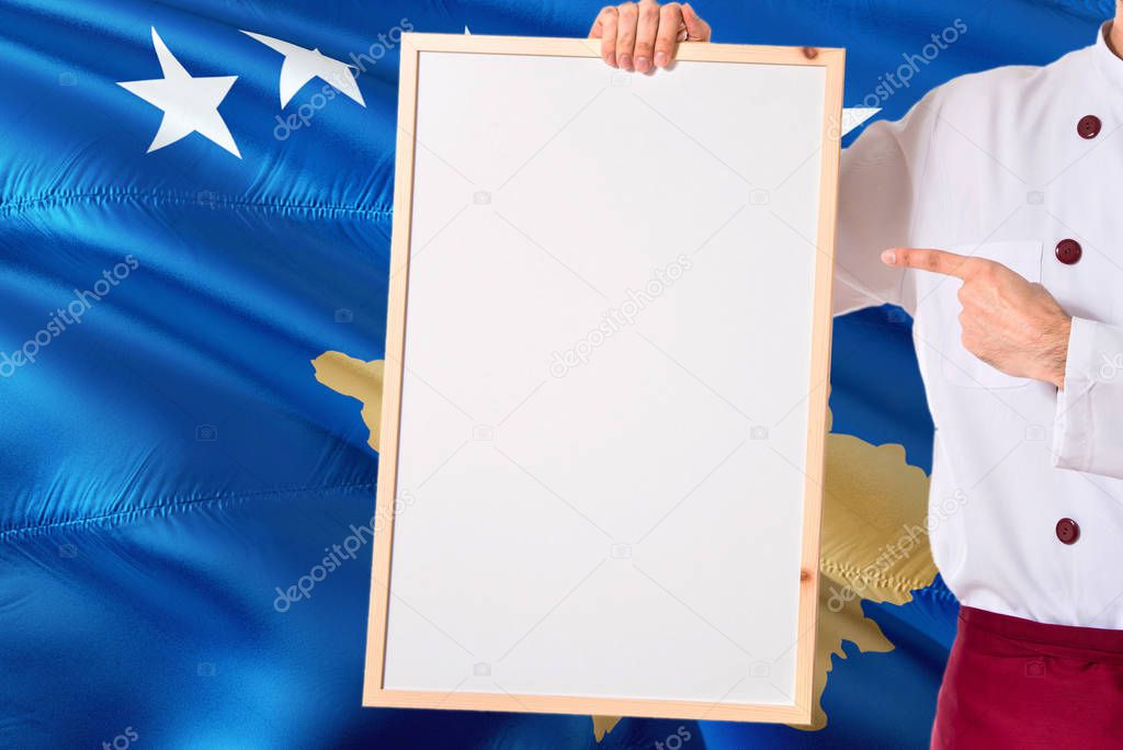 Chef holding blank whiteboard menu on Kosovo flag background. Cook wearing uniform pointing space for text.