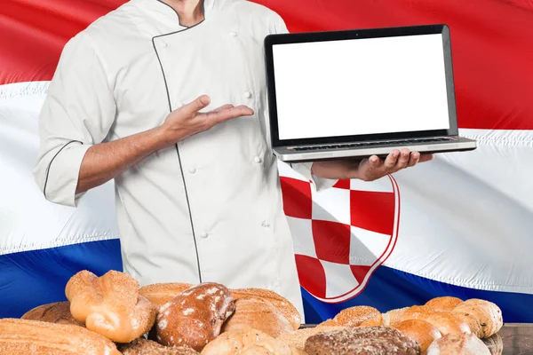 克罗地亚贝克拿着笔记本电脑在克罗地亚旗子和面包背景 厨师穿着统一的指向空白屏幕的复制空间 — 图库照片