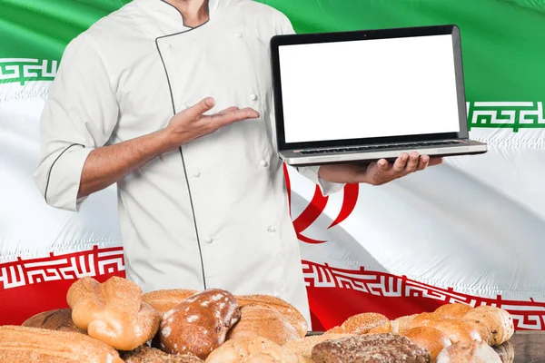 Iranian Baker holding laptop on Iran flag and breads background. Chef wearing uniform pointing blank screen for copy space.