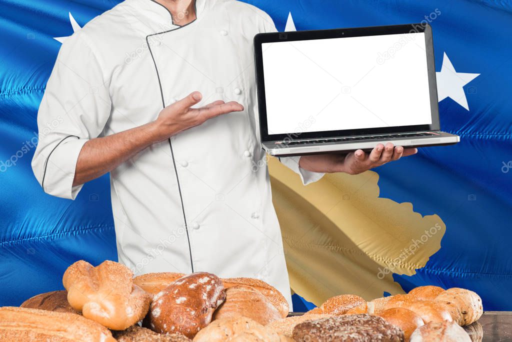 Baker holding laptop on Kosovo flag and breads background. Chef wearing uniform pointing blank screen for copy space.