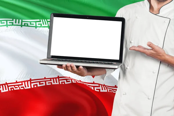 Iranian Chef holding laptop with blank screen on Iran flag background. Cook wearing uniform and pointing laptop for copy space.