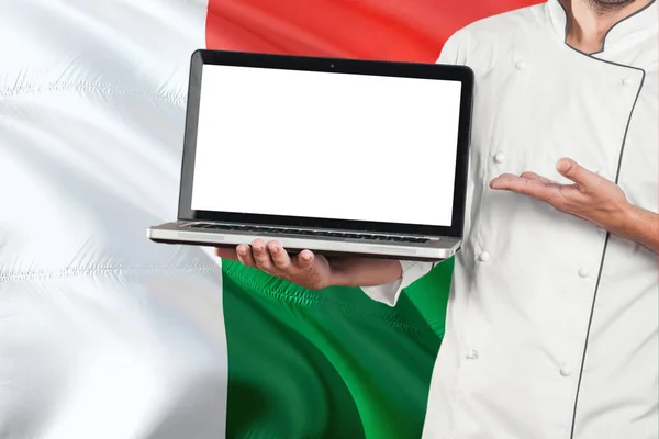 マダガスカルの旗の背景に空白の画面を持つノート パソコンを保持しているマダガスカル シェフ コピー スペースの制服とポインティングのラップトップを着て調理します — ストック写真