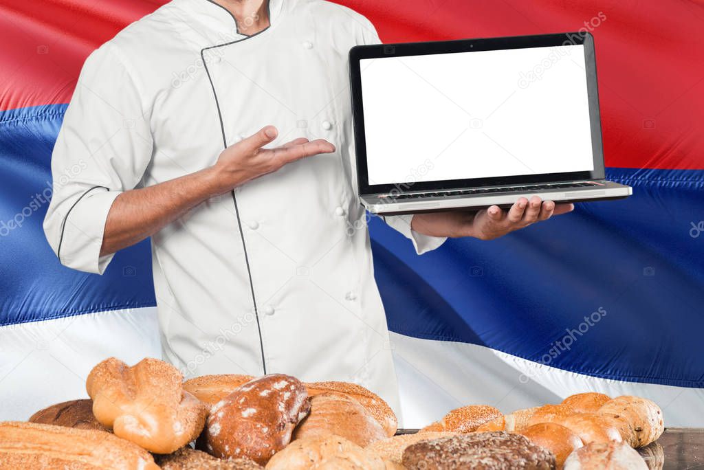 Serbian Baker holding laptop on Serbia flag and breads background. Chef wearing uniform pointing blank screen for copy space.