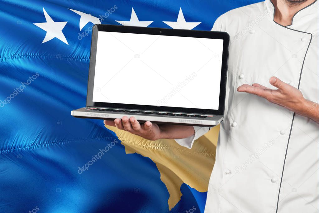 Chef holding laptop with blank screen on Kosovo flag background. Cook wearing uniform and pointing laptop for copy space.