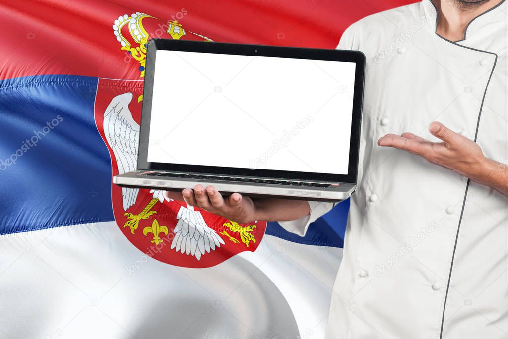 Serbian Chef holding laptop with blank screen on Serbia flag background. Cook wearing uniform and pointing laptop for copy space.