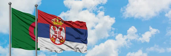 Bandera de Argelia y Serbia ondeando en el viento contra el cielo azul nublado blanco juntos. Concepto diplomático, relaciones internacionales . — Foto de Stock