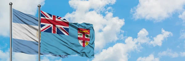 Bandera de Argentina y Fiyi ondeando en el viento contra el cielo azul nublado blanco juntos. Concepto diplomático, relaciones internacionales . — Foto de Stock
