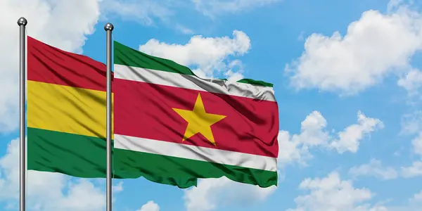 Bandera de Bolivia y Surinam ondeando en el viento contra el cielo azul nublado blanco juntos. Concepto diplomático, relaciones internacionales . — Foto de Stock