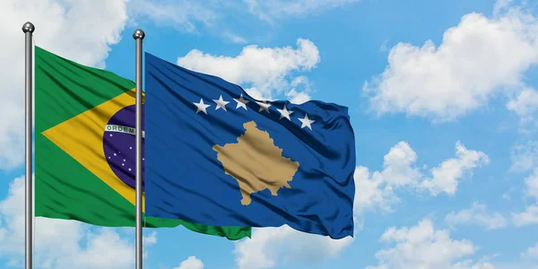 O Brasil e a bandeira do Kosovo agitando no vento contra o céu azul nublado branco juntos. Conceito de diplomacia, relações internacionais . — Fotografia de Stock