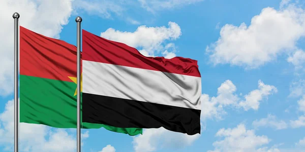 Burkina Faso y la bandera de Yemen ondeando en el viento contra el cielo azul nublado blanco juntos. Concepto diplomático, relaciones internacionales . — Foto de Stock