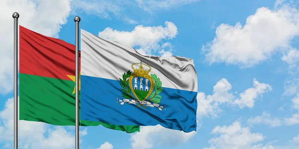 Burkina Faso y la bandera de San Marino ondeando en el viento contra el cielo azul nublado blanco juntos. Concepto diplomático, relaciones internacionales . — Foto de Stock