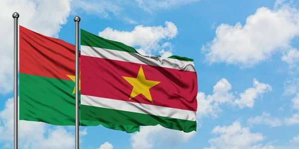 Burkina Faso y la bandera de Surinam ondeando en el viento contra el cielo azul nublado blanco juntos. Concepto diplomático, relaciones internacionales . — Foto de Stock