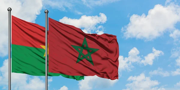 Burkina Faso y la bandera de Marruecos ondeando en el viento contra el cielo azul nublado blanco juntos. Concepto diplomático, relaciones internacionales . — Foto de Stock