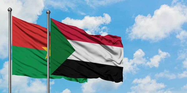 Burkina Faso y la bandera de Sudán ondeando en el viento contra el cielo azul nublado blanco juntos. Concepto diplomático, relaciones internacionales . — Foto de Stock