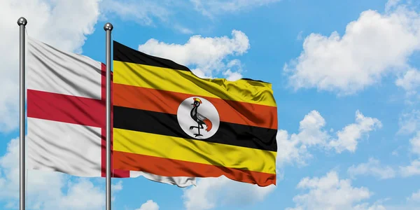 A bandeira da Inglaterra e Uganda agitando no vento contra o céu azul nublado branco juntos. Conceito de diplomacia, relações internacionais . — Fotografia de Stock