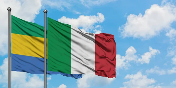 Bandera de Gabón e Italia ondeando en el viento contra el cielo azul nublado blanco juntos. Concepto diplomático, relaciones internacionales . — Foto de Stock