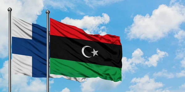 Bandera de Finlandia y Libia ondeando en el viento contra el cielo azul nublado blanco juntos. Concepto diplomático, relaciones internacionales . — Foto de Stock