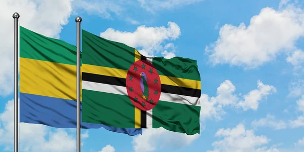 Gabão e Dominica bandeira acenando no vento contra o céu azul nublado branco juntos. Conceito de diplomacia, relações internacionais . — Fotografia de Stock