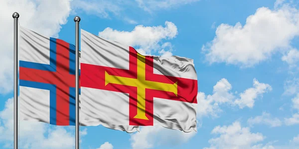 Islas Feroe y la bandera de Guernsey ondeando en el viento contra el cielo azul nublado blanco juntos. Concepto diplomático, relaciones internacionales . — Foto de Stock