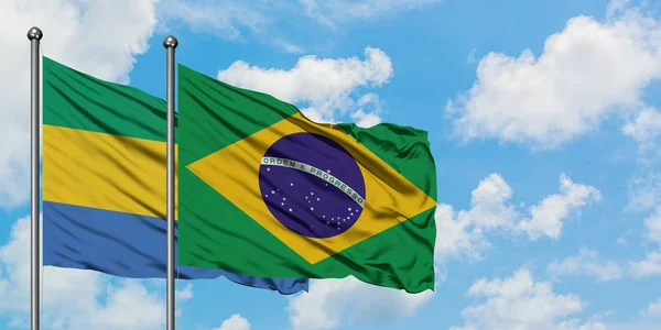 Gabón y la bandera de Brasil ondeando en el viento contra el cielo azul nublado blanco juntos. Concepto diplomático, relaciones internacionales . — Foto de Stock