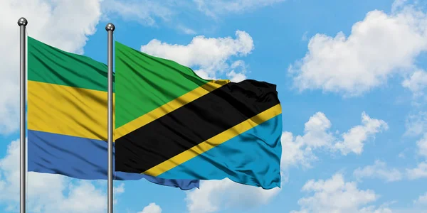 Gabón y la bandera de Tanzania ondeando en el viento contra el cielo azul nublado blanco juntos. Concepto diplomático, relaciones internacionales . — Foto de Stock