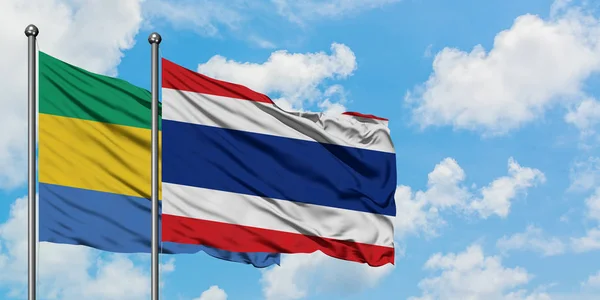 Gabón y la bandera de Tailandia ondeando en el viento contra el cielo azul nublado blanco juntos. Concepto diplomático, relaciones internacionales . — Foto de Stock