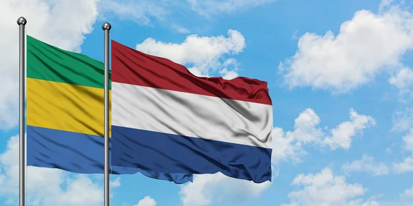 Gabón y la bandera de los Países Bajos ondeando en el viento contra el cielo azul nublado blanco juntos. Concepto diplomático, relaciones internacionales . — Foto de Stock