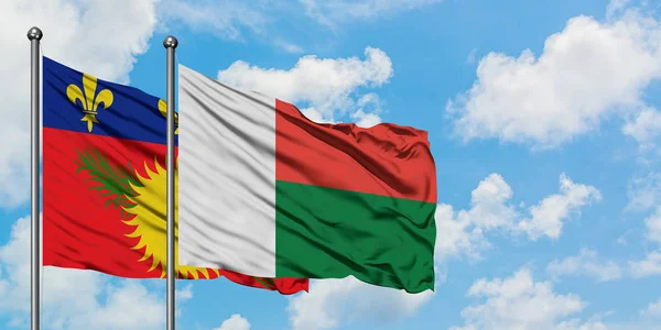 Guadalupe y la bandera de Madagascar ondeando en el viento contra el cielo azul nublado blanco juntos. Concepto diplomático, relaciones internacionales . — Foto de Stock