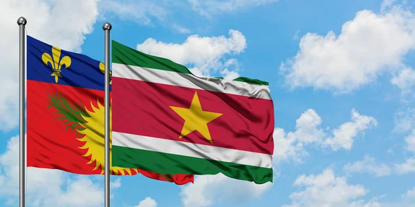 Guadalupe y la bandera de Surinam ondeando en el viento contra el cielo azul nublado blanco juntos. Concepto diplomático, relaciones internacionales . — Foto de Stock