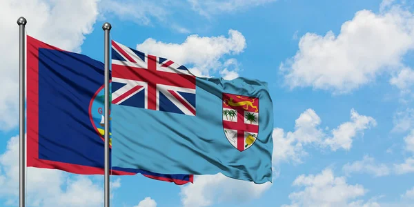 Bandera de Guam y Fiyi ondeando en el viento contra el cielo azul nublado blanco juntos. Concepto diplomático, relaciones internacionales . — Foto de Stock