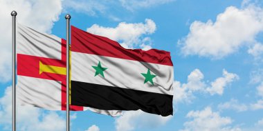 Guernsey ve Suriye bayrağı birlikte beyaz bulutlu mavi gökyüzüne karşı rüzgarda sallayarak. Diplomasi kavramı, uluslararası ilişkiler.