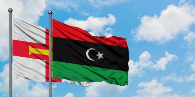 Guernsey ve Libya bayrağı birlikte beyaz bulutlu mavi gökyüzüne karşı rüzgarda sallayarak. Diplomasi kavramı, uluslararası ilişkiler.