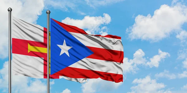 Guernsey y la bandera de Puerto Rico ondeando en el viento contra el cielo azul nublado blanco juntos. Concepto diplomático, relaciones internacionales . — Foto de Stock