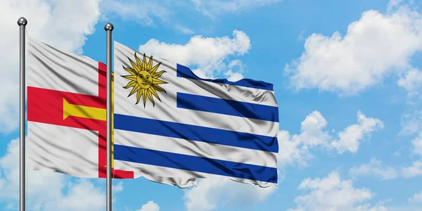 Bandera de Guernsey y Uruguay ondeando en el viento contra el cielo azul nublado blanco juntos. Concepto diplomático, relaciones internacionales . —  Fotos de Stock