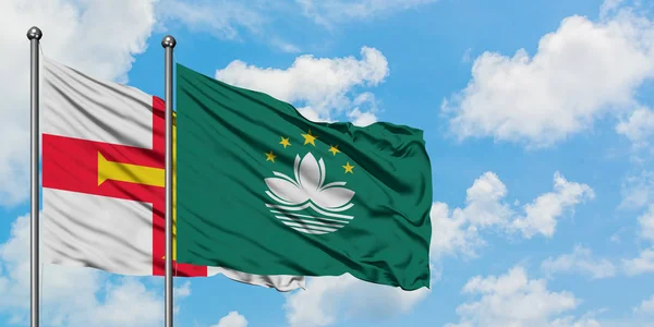 Bandera de Guernsey y Macao ondeando en el viento contra el cielo azul nublado blanco juntos. Concepto diplomático, relaciones internacionales . — Foto de Stock