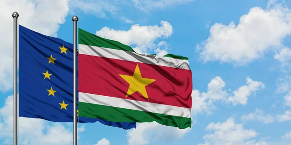 Bandera de la Unión Europea y Surinam ondeando en el viento contra el cielo azul nublado blanco juntos. Concepto diplomático, relaciones internacionales . — Foto de Stock