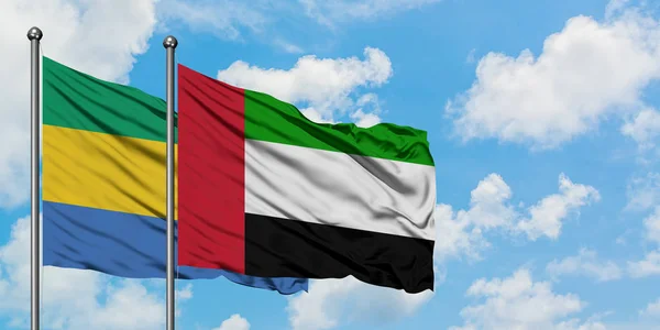 Gabón y la bandera de los Emiratos Árabes Unidos ondeando en el viento contra el cielo azul nublado blanco juntos. Concepto diplomático, relaciones internacionales . — Foto de Stock