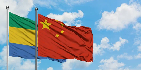 Gabon og Kina flag vinker i vinden mod hvid overskyet blå himmel sammen. Diplomati koncept, internationale relationer . - Stock-foto
