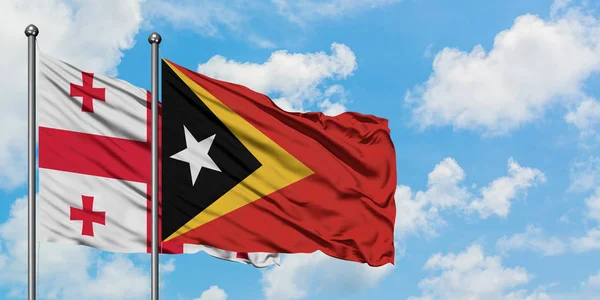 Bandera de Georgia y Timor Oriental ondeando en el viento contra el cielo azul nublado blanco juntos. Concepto diplomático, relaciones internacionales . — Foto de Stock