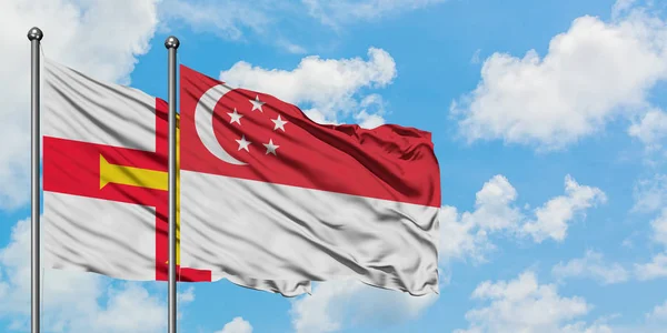 Guernsey e a bandeira de Singapura agitando no vento contra o céu azul nublado branco juntos. Conceito de diplomacia, relações internacionais . — Fotografia de Stock