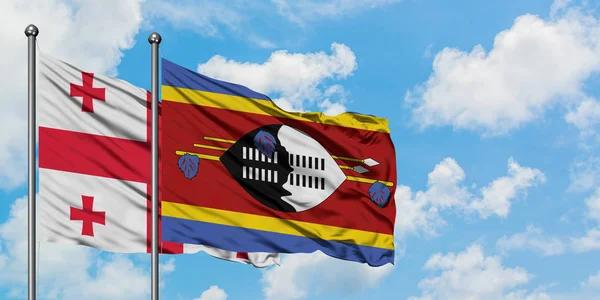 Bandera de Georgia y Suazilandia ondeando en el viento contra el cielo azul nublado blanco juntos. Concepto diplomático, relaciones internacionales . — Foto de Stock